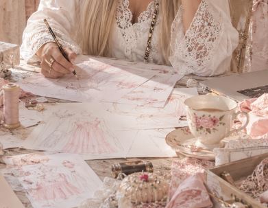 Designer sketching pink nightgown illustrations at a lace-covered desk with a floral teacup
